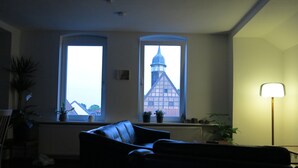 Sala de estar com sofá, duas janelas com vista para um edifício, luminária de chão e radiador.