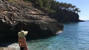 Eine Frau in Bikini und Hut steht am Rand eines felsigen Strandes, im Hintergrund klares Wasser und Bäume.