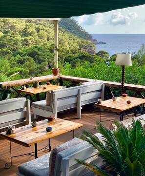 An outdoor seating area with wooden tables and benches, surrounded by lush greenery and overlooking a coastal landscape. - Turan Hill Lounge (Fethiye)