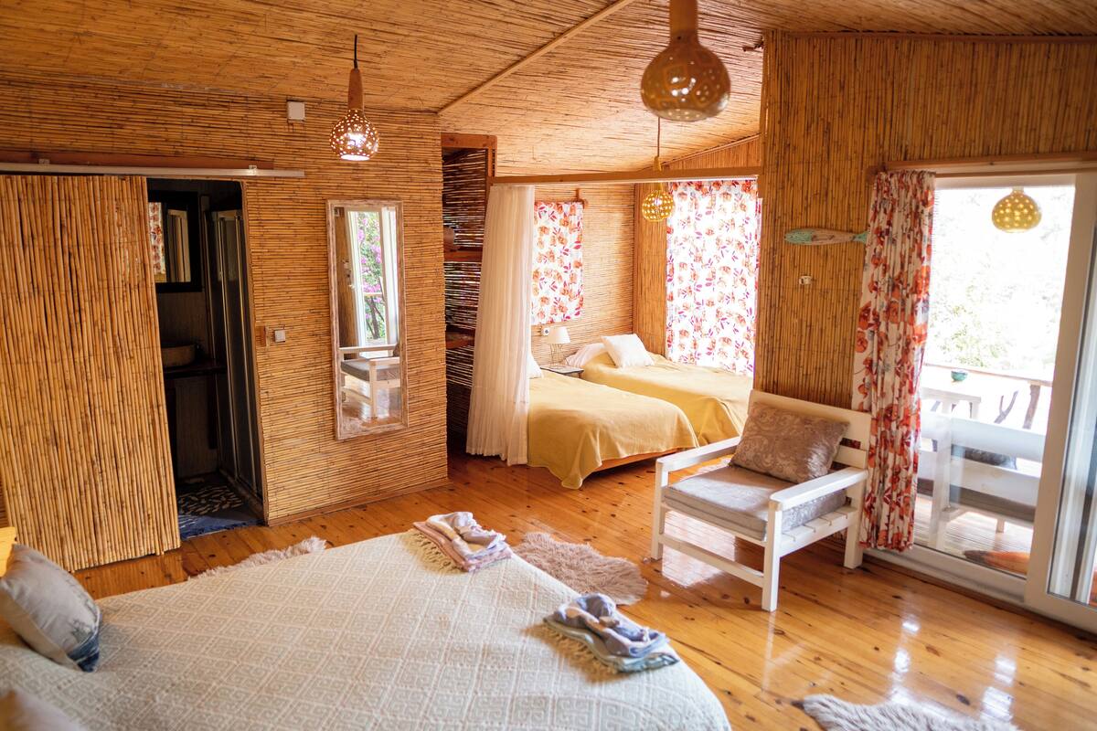 A wooden interior with a bed, two bedside tables, a chair, and a sliding glass door to an outdoor area.
