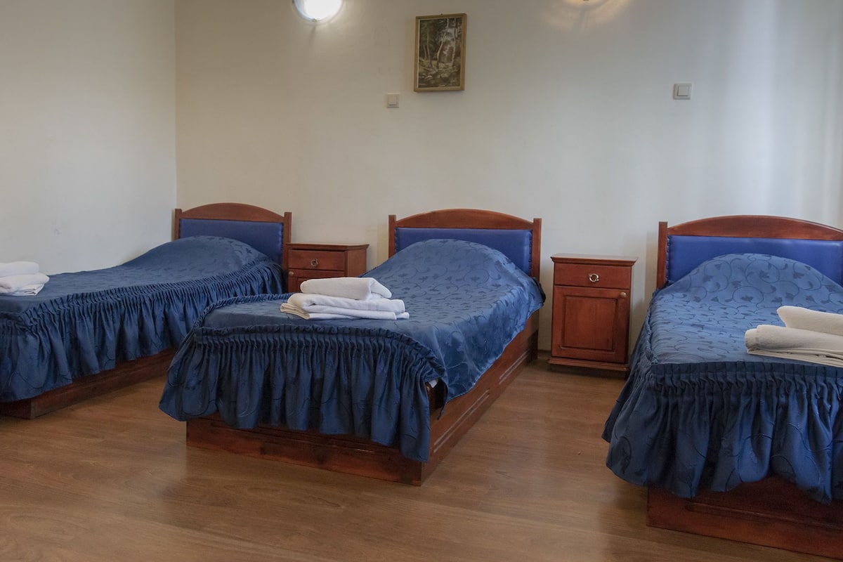 A room with three wooden beds, each with a blue bedspread and white towels. There are wooden nightstands between the beds, and a small wooden dresser to the right.