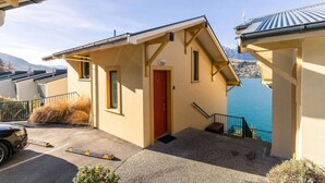 Ein kleines, eingeschossiges Gebäude mit roter Tür, einer Treppe, die zu einem Balkon führt, und Blick auf ein Gewässer sowie Berge im Hintergrund.