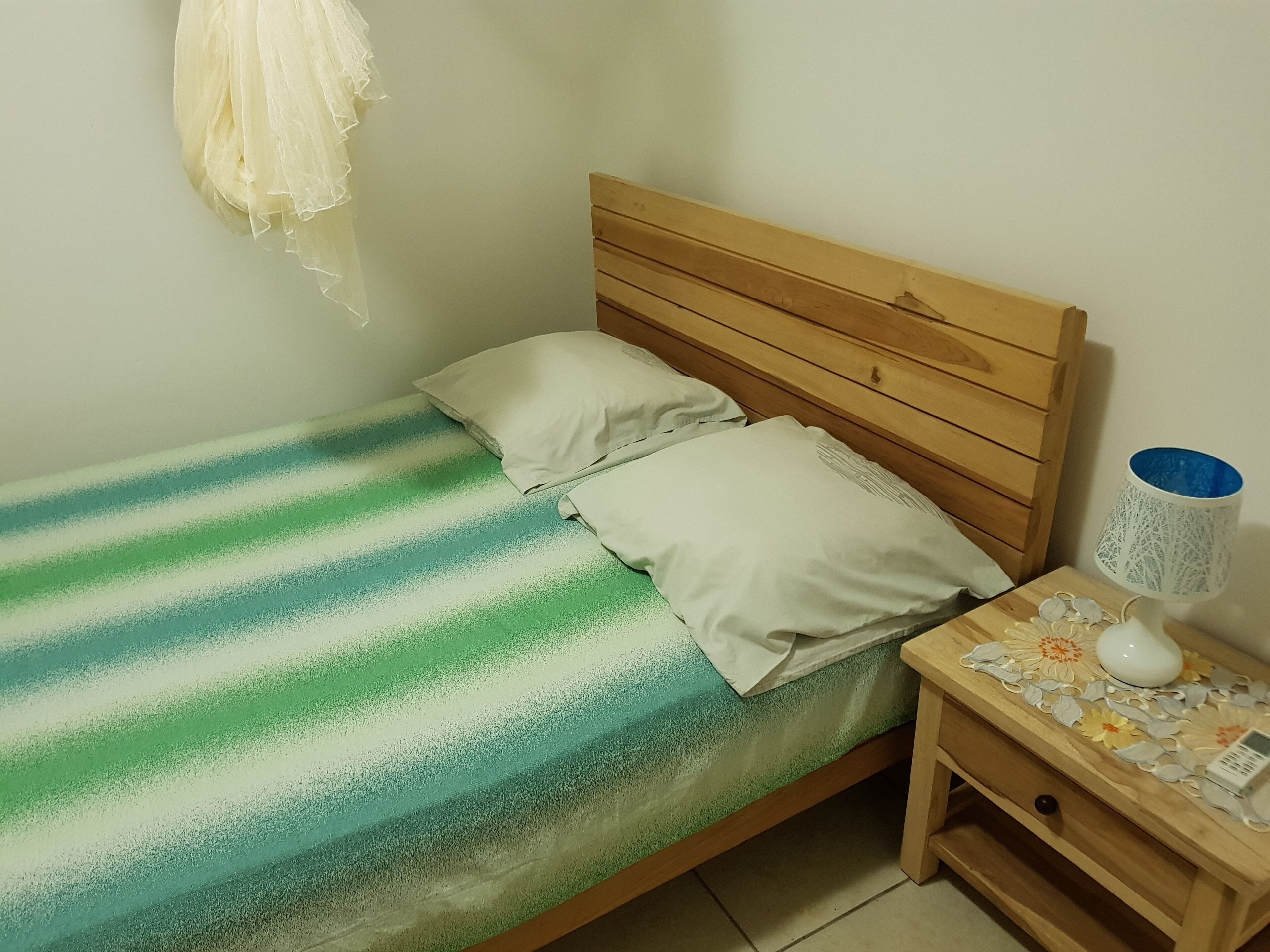 Ein Einzelbett mit gestreifter Bettdecke, zwei Kissen, ein hölzernes Kopfbrett, ein Nachttisch mit Lampe und ein an der Wand befestigtes Element.
