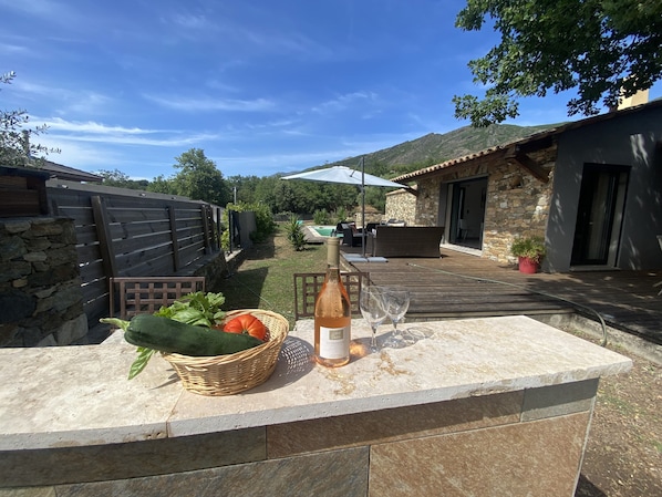 A patio area with a stone countertop, a wicker basket with vegetables, a bottle of wine, and two glasses.