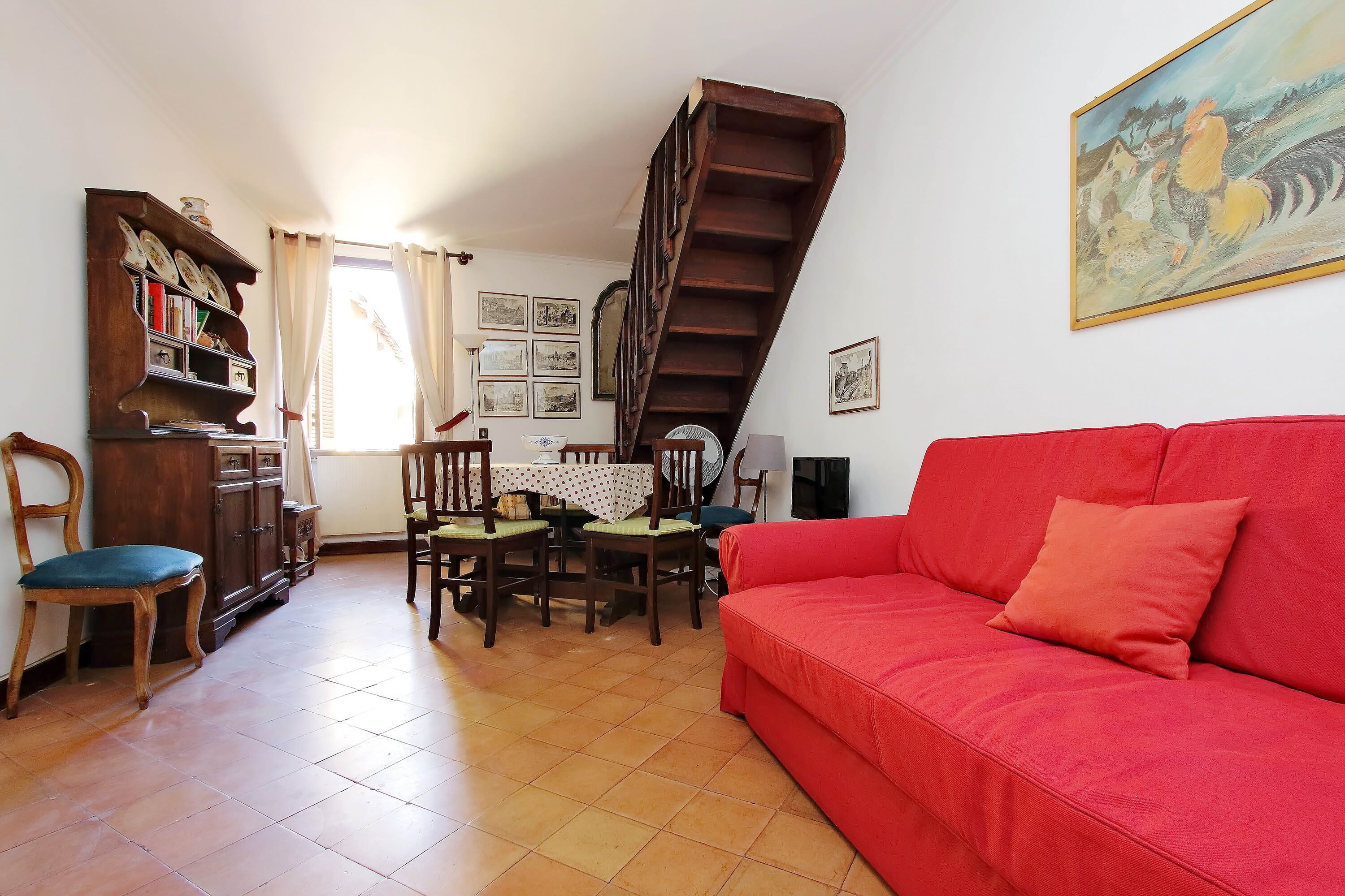 A living room with a red sofa, wooden dining table, chairs, a staircase, and a bookshelf.