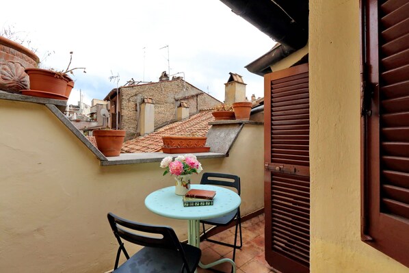 A small outdoor seating area with a table and chairs, a potted plant, and a view of a residential street.