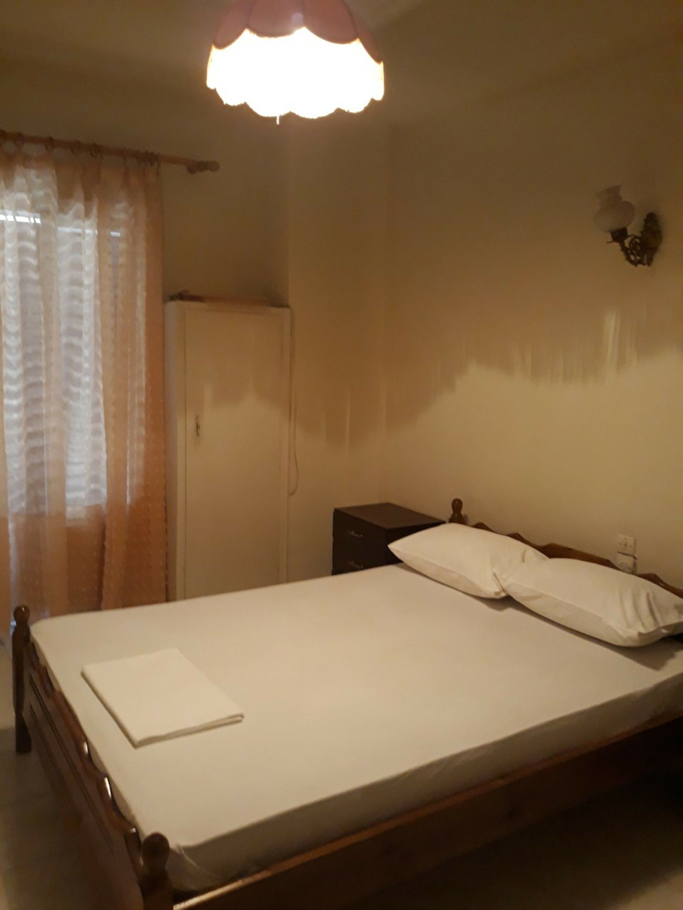 Una cama individual con colchón y almohadas blancos, cabecera de madera, mesita de noche con lámpara y una puerta cerrada.