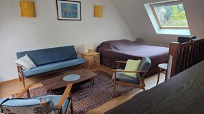Ein Schlafzimmer im Loft-Stil mit schrägverdeckter Decke, einem blauen Sofa, einem Holzstuhl, einem kleinen Tisch, einem Bett mit dunkler Bettdecke und einem Fenster mit Blick auf Bäume.