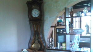 A dining area with a wooden table and chairs, a large wall clock, and a kitchen area with shelves and appliances.