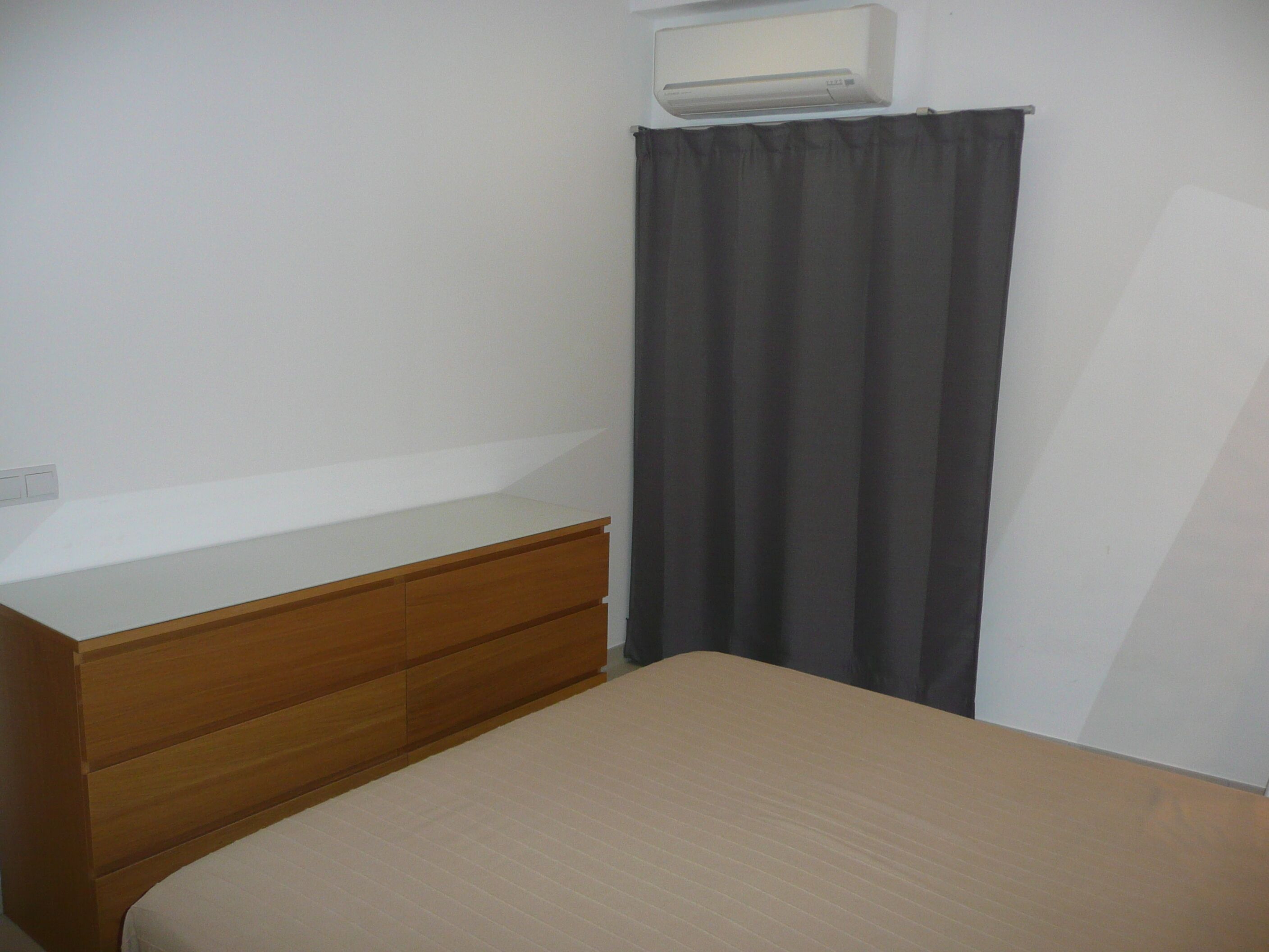Une chambre à coucher avec un lit, une commode en bois, un rideau foncé et un appareil de climatisation fixé au mur.