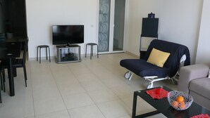 A modern living room with a flat-screen TV, a sofa, a dining table with chairs, and a sliding glass door leading to a balcony.