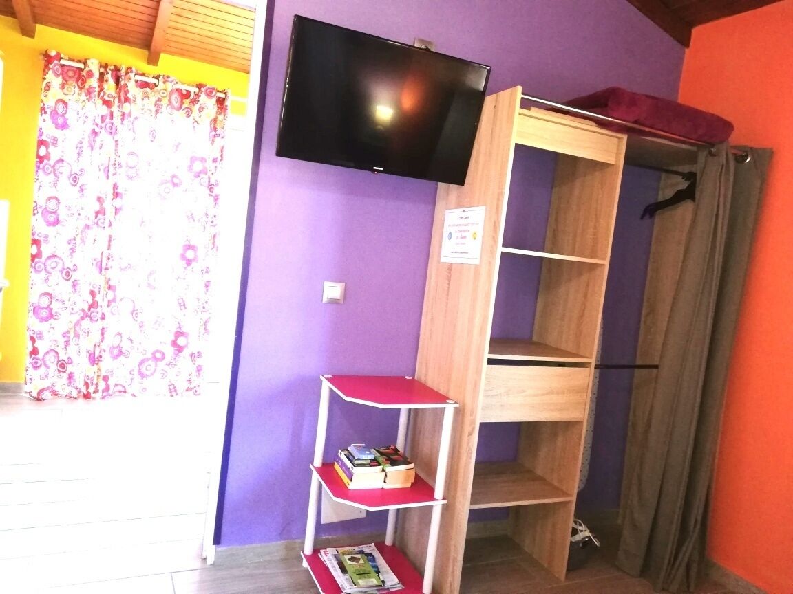A room with a TV mounted on the wall, a wooden shelf unit with books, and a window with floral curtains.