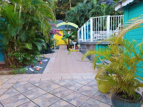 A pathway leading to a building with a yellow facade, surrounded by lush greenery and a tiled floor.