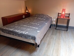 Una cama bien hecha con una colcha gris con frases motivacionales, una mesita de noche con una lámpara y una pequeña mesita con una lámpara roja.