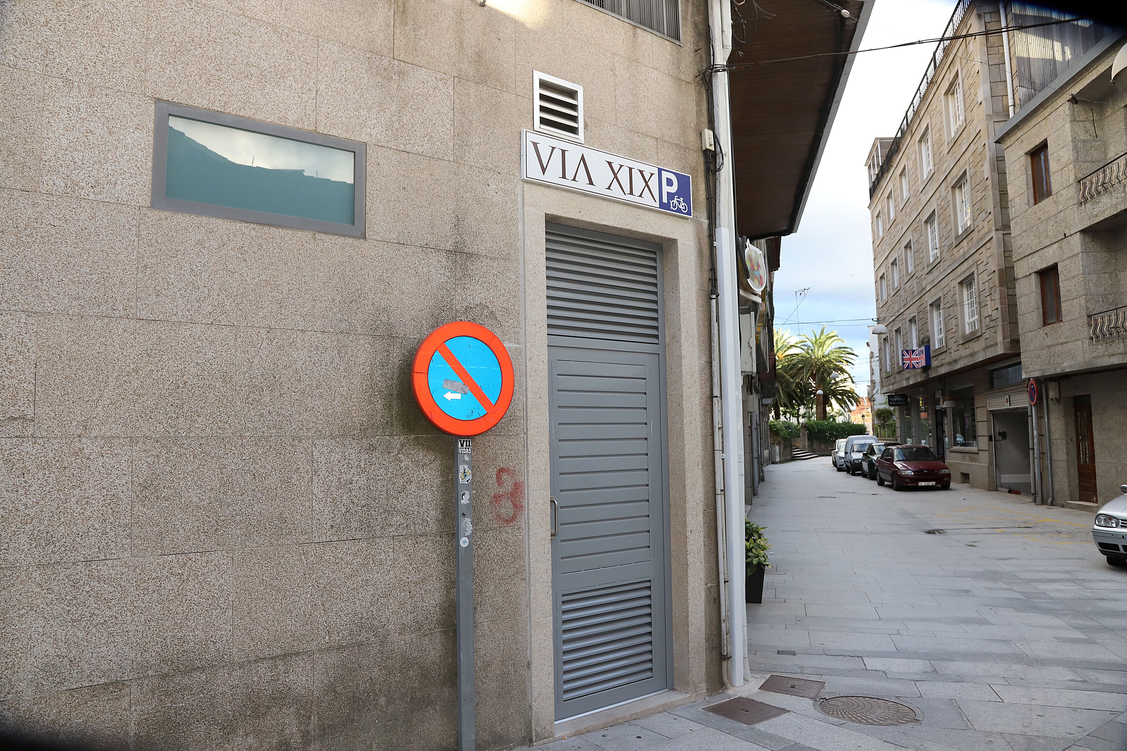 A street with a no parking sign, a building with a 'VIA XIX' sign, and parked cars.