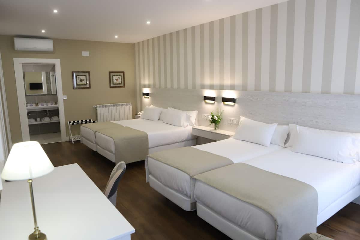 A hotel room with two beds, a desk, a chair, a lamp, a radiator, and framed pictures on the wall.
