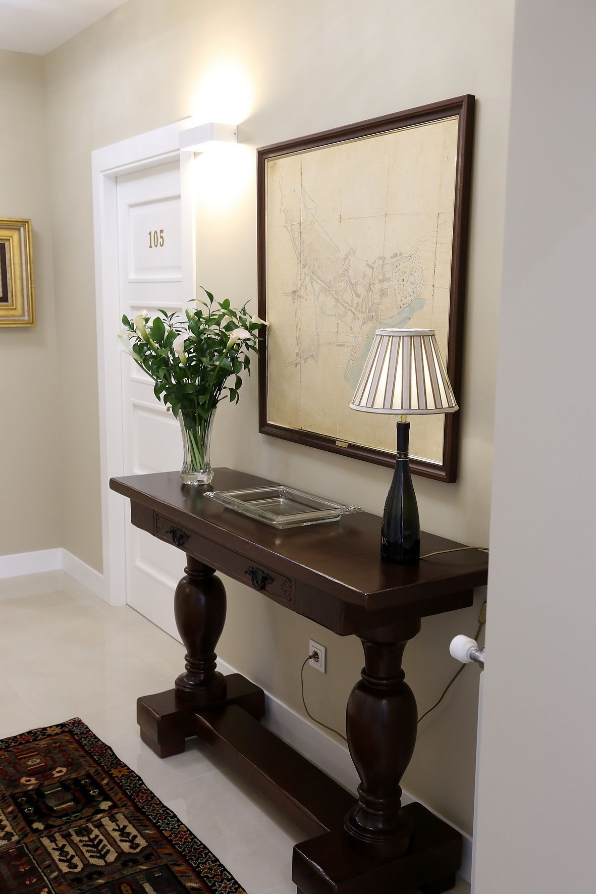 A dark wooden console table with a lamp, a vase with white flowers, and a framed map on the wall.