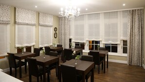 Un comedor con varias mesas y sillas, una araña y ventanas amplias con cortinas.