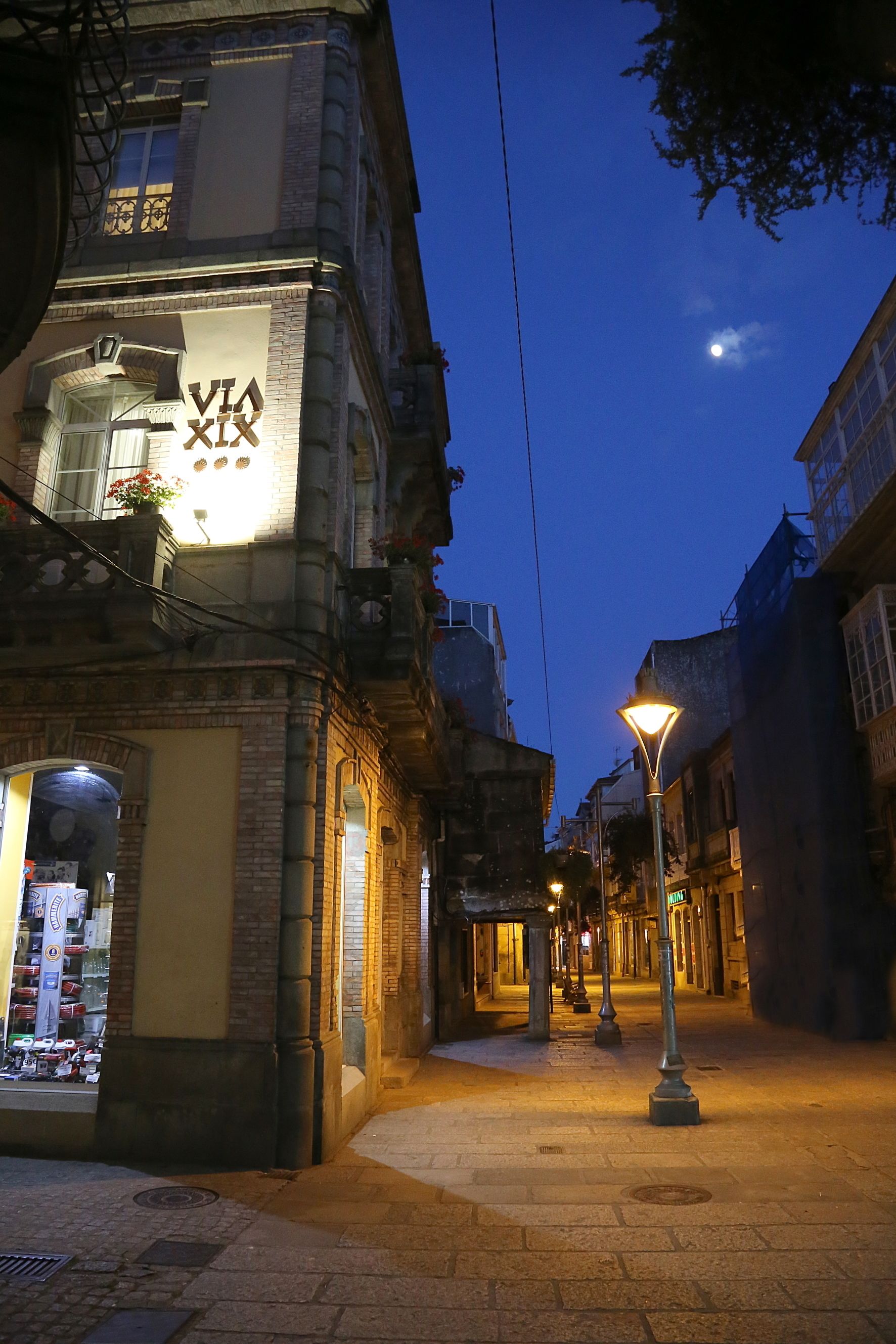 A street at dusk with a building featuring a 'VIA XIX' sign.