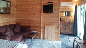 Ein gemütliches Cabin-Interieur mit Holzvertäfelung, einer Couch, einem kleinen Tisch, einem an der Wand befestigten Fernseher und einer Küchenzeile mit Schränken und einem Fenster.