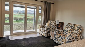 Ein Hotelzimmer mit einer großen Schiebeglasdoor zu einer ländlichen Aussicht, zwei Armlehnstühle mit Blumenmuster, ein kleiner Tisch mit Lampe und ein Bett mit gemustertem Überwurf.