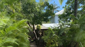 Un sentier en bois mène à un bâtiment rustique, entouré d’une végétation luxuriante et de petits galets blancs.