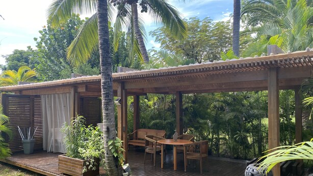 Une terrasse en bois dotée d’un espace salon abrité, entourée d’une végétation luxuriante et de palmiers.