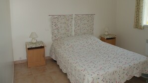 Un dormitorio con una cama, dos mesitas de noche, una mesita auxiliar, un cuadro en la pared y una ventana con cortinas.