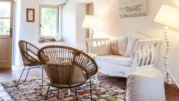 Un accogliente soggiorno con soffitto alto, un divano bianco, due poltrone in vimini, un tappeto colorato e una lampada bianca.