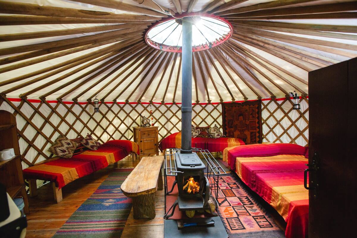 A yurt with a central wood-burning stove, two beds with red bedding, a wooden bench, and a patterned rug.