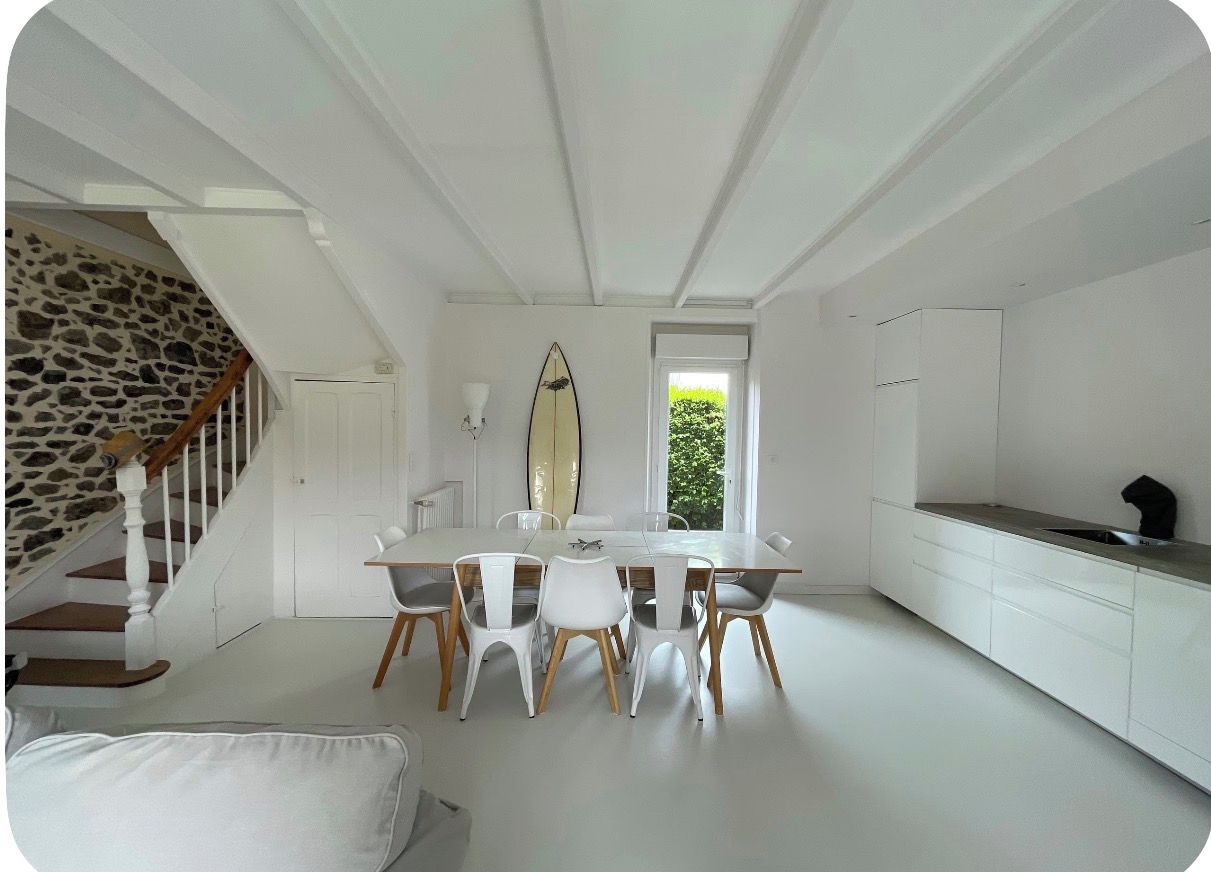 A modern interior with a dining area, white furniture, a surfboard, and a staircase with a stone wall.
