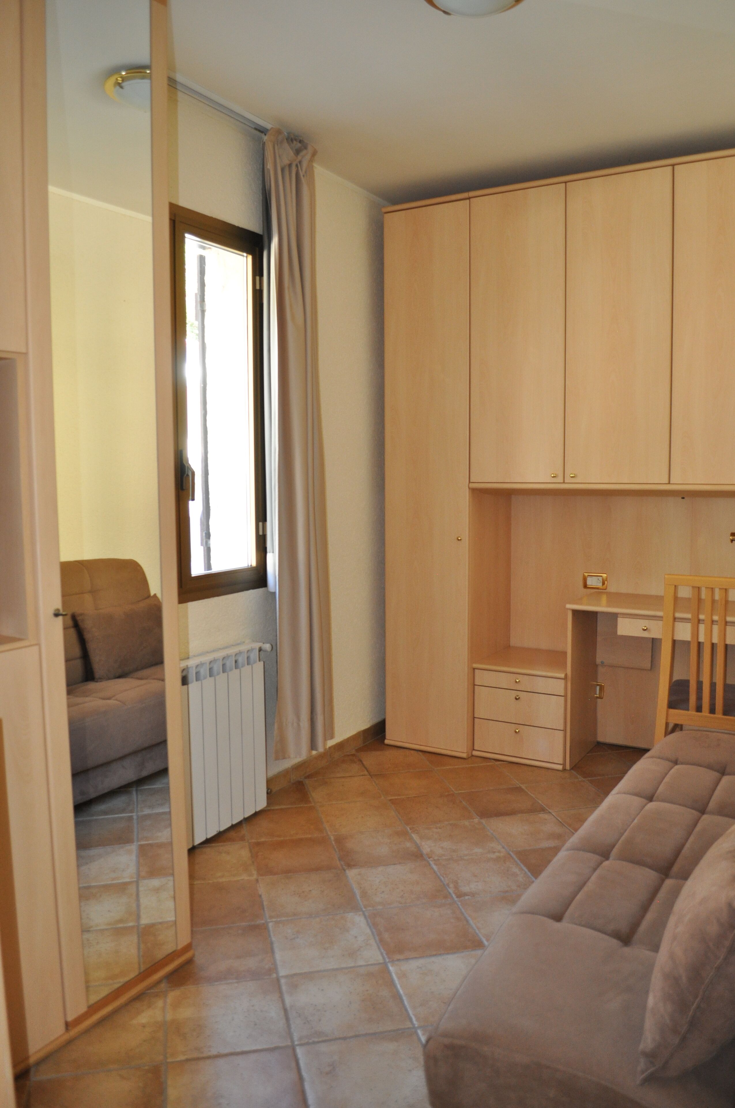 Een compacte kamer met een bank, een klein bureau en een stoel. Een raam met gordijnen is zichtbaar, en een radiator hangt aan de muur.
