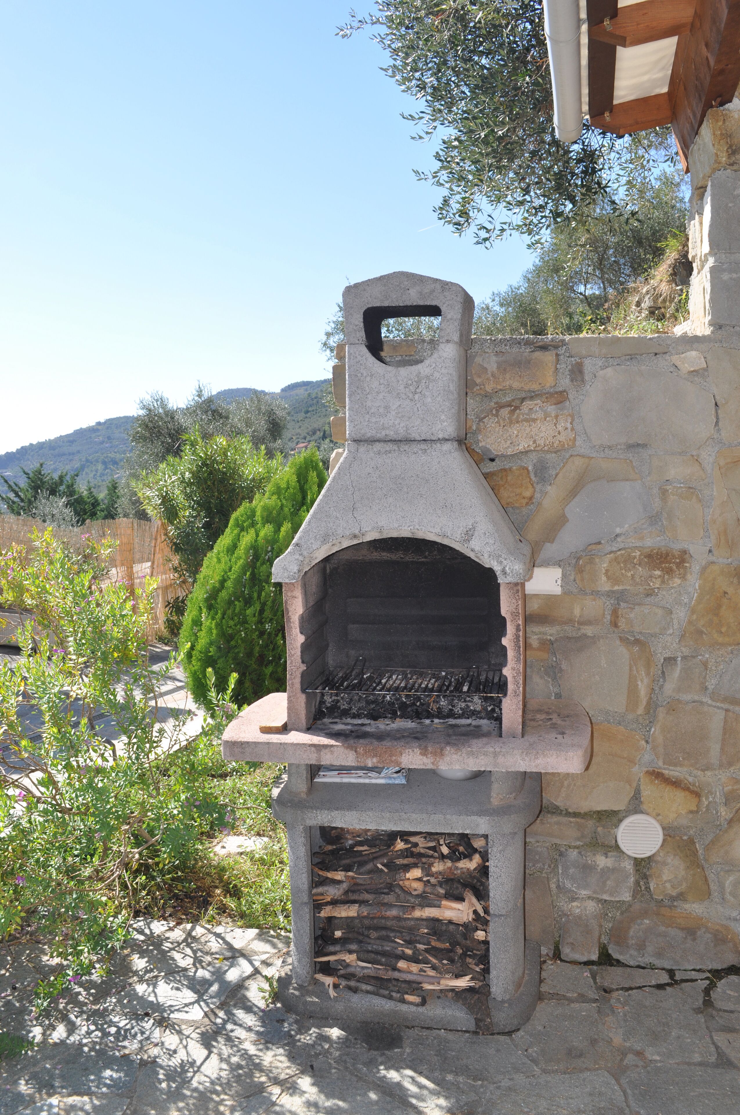 Un four à pizza extérieur en pierre, doté d’un foyer, adossé à un mur de pierre et entouré de verdure.