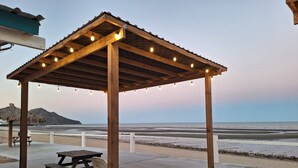 Een houten paviljoen aan het strand met lichtjes, een picknicktafel en stoelen.