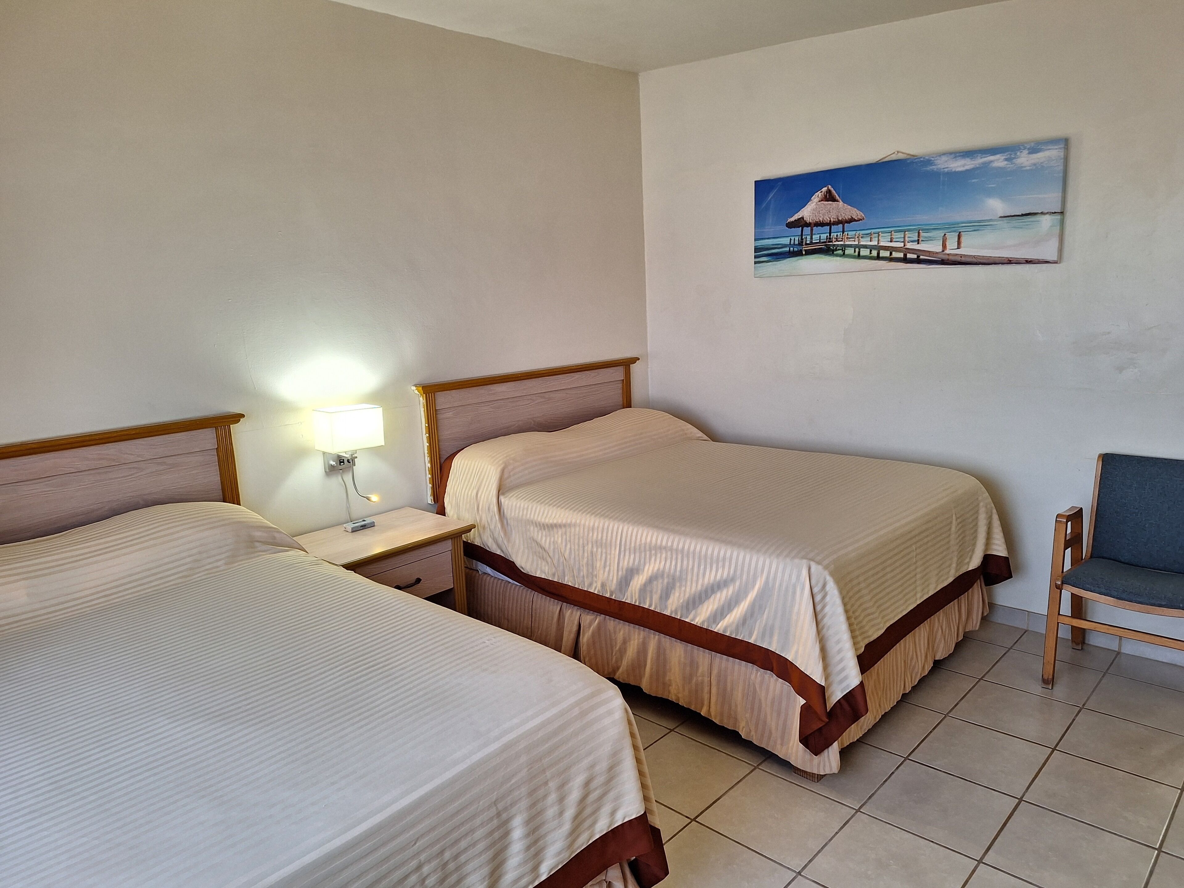 A hotel room with two beds, a nightstand with a lamp, a chair, and a painting of a beach scene on the wall.