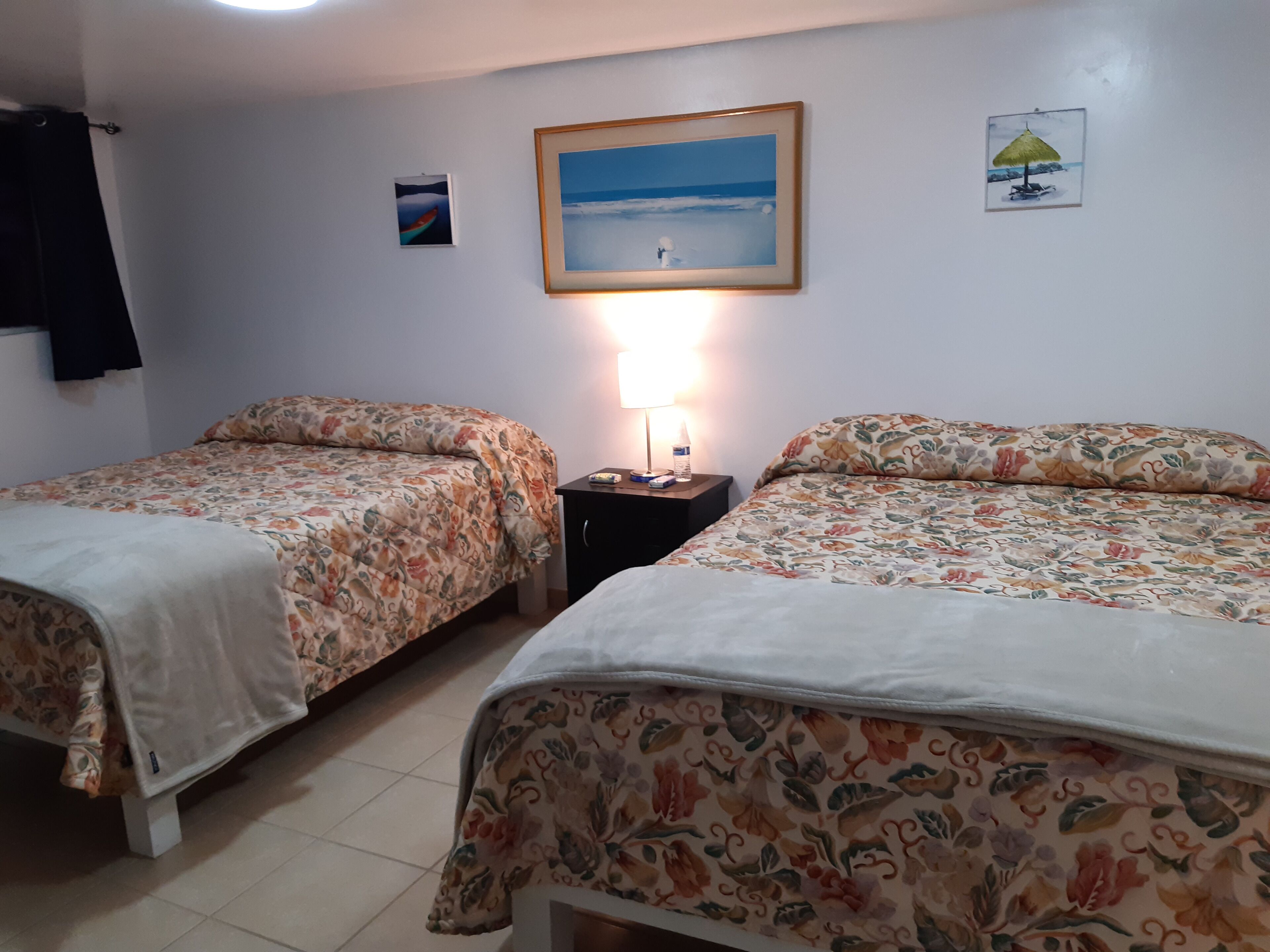 Une chambre avec deux lits, une table de chevet avec une lampe et un tableau représentant une scène de plage.