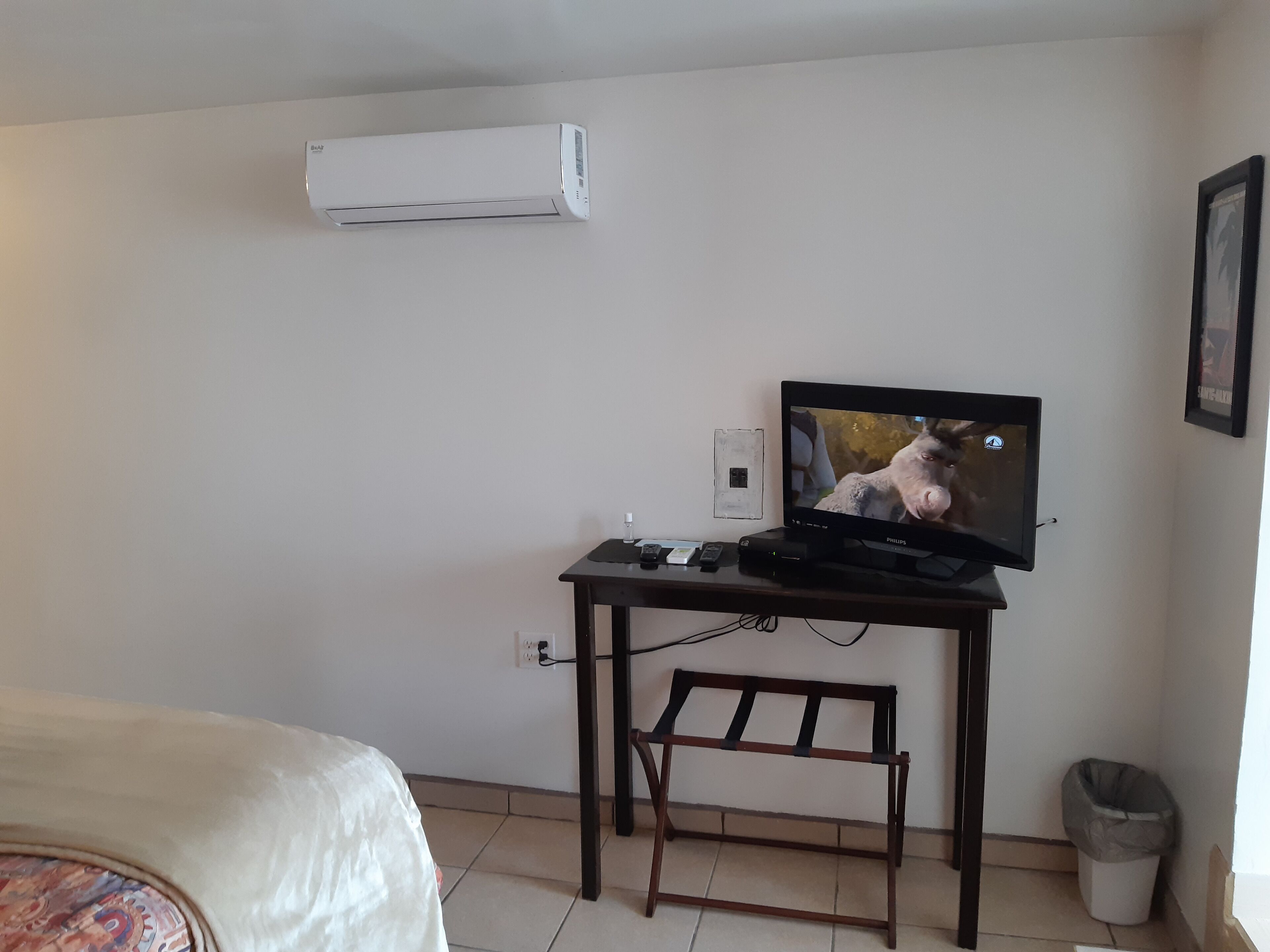 Une petite pièce comprenant un lit, un bureau avec une télévision, un climatiseur et une poubelle.