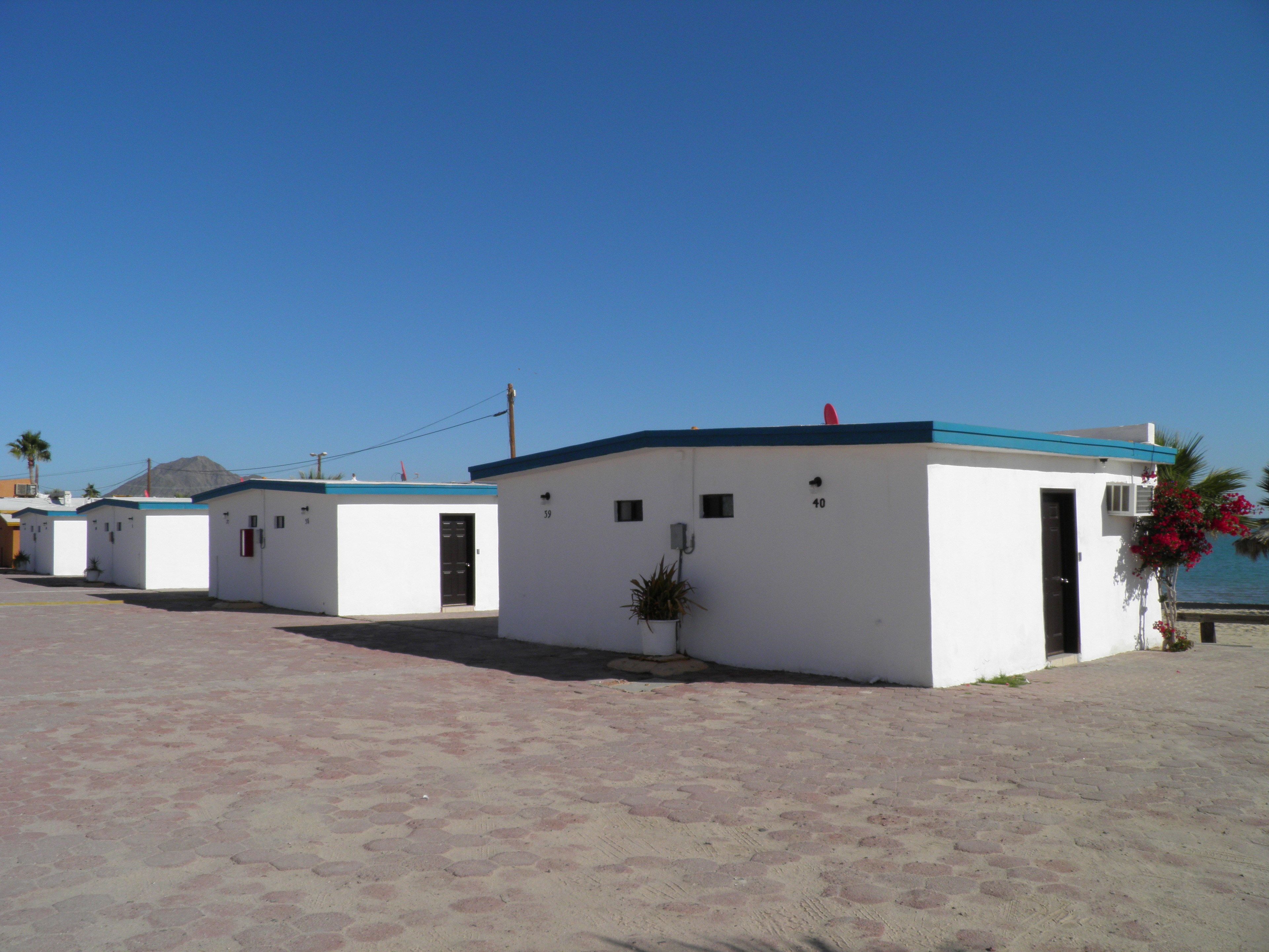 Una hilera de edificios pequeños y blancos con techos azules, con los números 39 y 40, sobre un terreno arenoso bajo un cielo azul despejado.