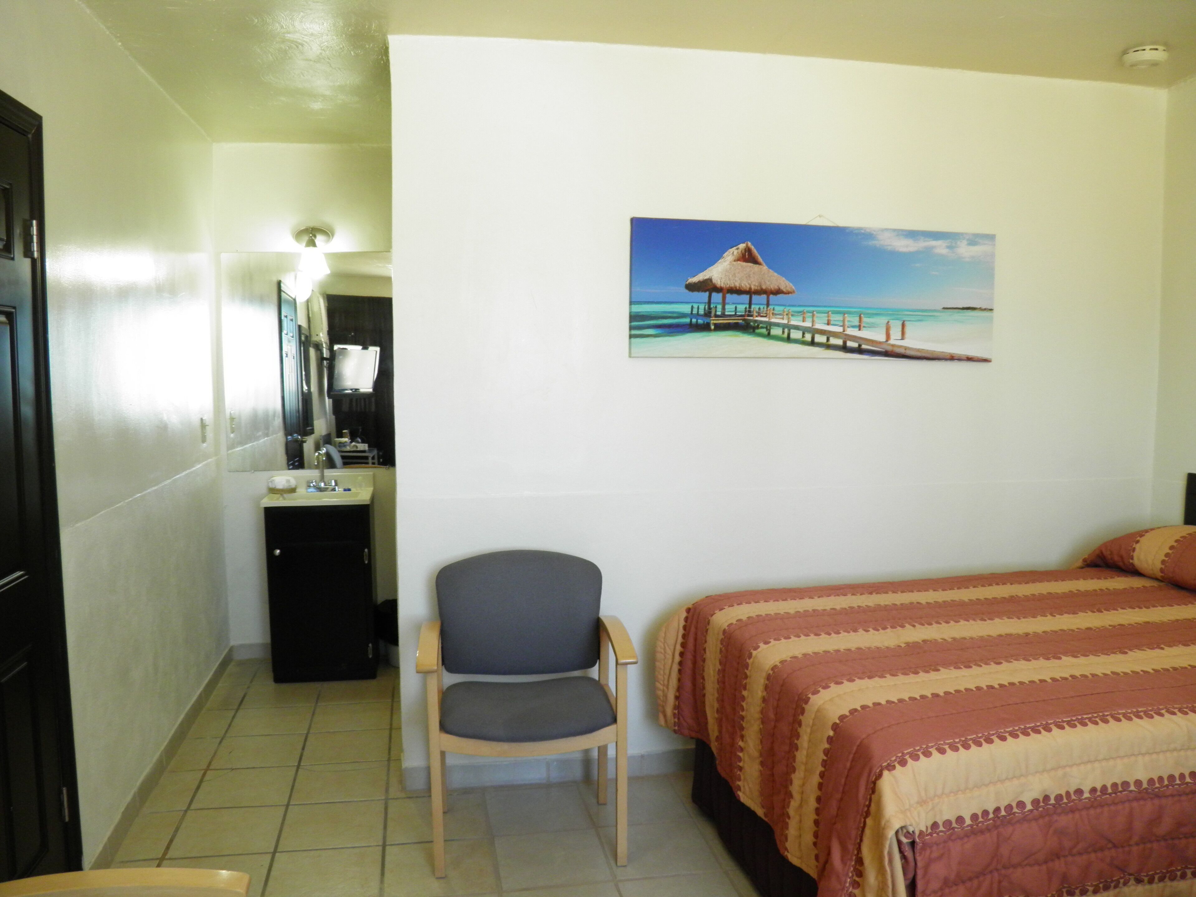 Quarto de hotel com cama, poltrona, escrivaninha com espelho e quadro de um cais na parede.