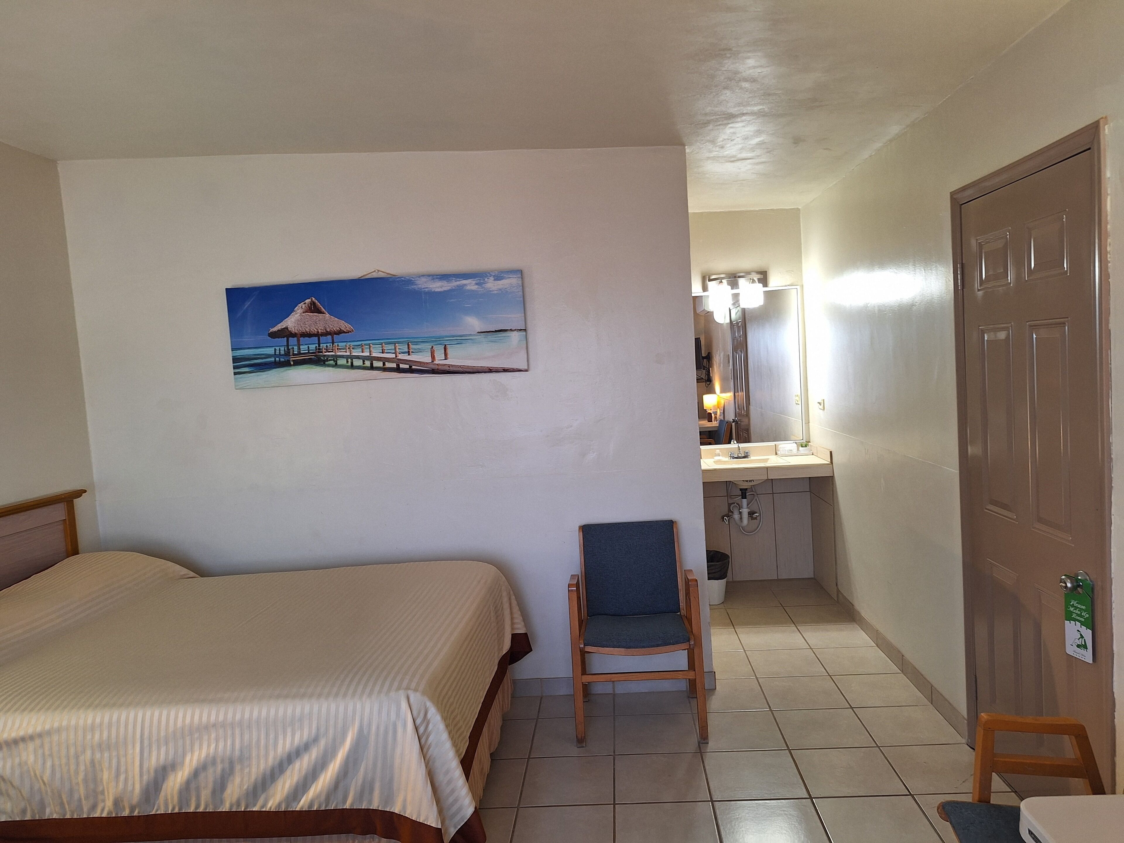 Habitación de hotel con cama, una silla, un cuadro de una playa, baño con espejo y puerta.