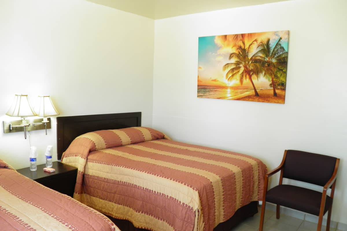 Une chambre d’hôtel avec deux lits, une chaise, une table de chevet avec des lampes et un tableau représentant un coucher de soleil sur des palmiers.