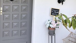 Una puerta gris con un aldabón metálico, un farol negro de pared, un cartel de bienvenida y una planta en maceta con flores.