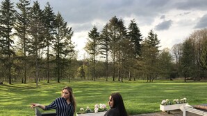 Dos personas sentadas en muebles de mimbre al aire libre en un espacio con césped, una valla blanca y árboles de fondo.