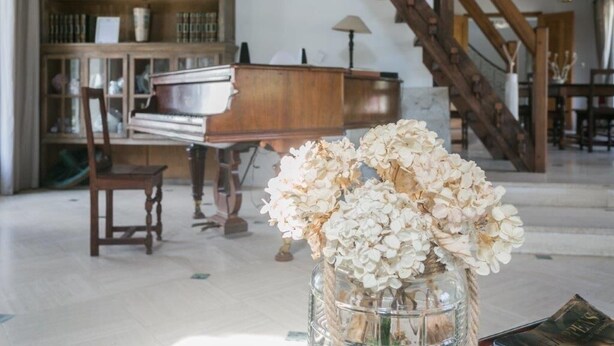 Una habitación con un piano, una silla, un florero con flores y una escalera.