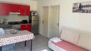 A compact kitchen with red cabinets, a microwave, and a refrigerator. There is a dining table with a floral tablecloth and a sofa with a red blanket.