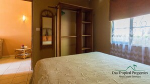 Quarto com cama, guarda-roupa, espelho, pequena mesa com livros e janela com vista.