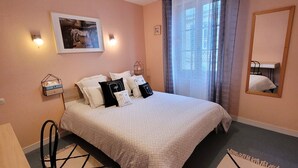 A neatly arranged bedroom with a bed, bedside tables, a chair, a hanging light fixture, a window with curtains, and a wall-mounted mirror. - Le Relais du Château (Plaine-et-Vallées)