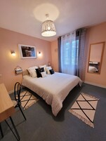 A neatly arranged bedroom with a bed, bedside tables, a chair, a hanging light fixture, a window with curtains, and a wall-mounted mirror.