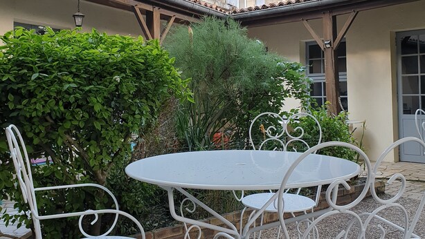 A courtyard with white metal tables and chairs, surrounded by greenery and a building with a tiled roof and shutters. - Le Relais du Château (Plaine-et-Vallées)