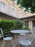 A courtyard with white metal tables and chairs, surrounded by greenery and a building with a tiled roof and shutters.